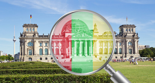 Das Reichstagsgebäude in Berlin, betrachtet durch eine Lupe, die in den Farben der Ampel-Koalition (Rot, Grün und Gelb) eingefärbt ist.