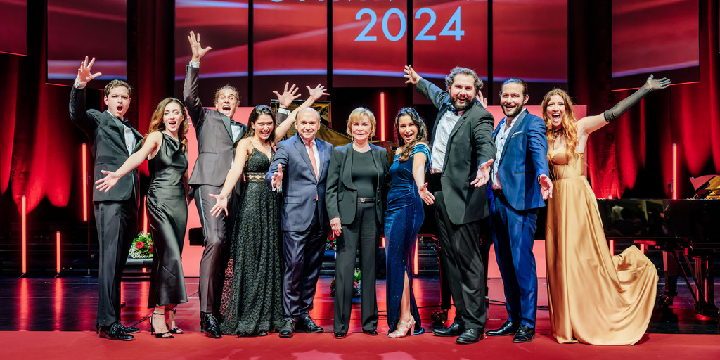 Gruppenfoto der Finalist:innen der NEUEN STIMMEN 2024 mit NEUE-STIMMEN-Gründerin Liz Mohn und dem Juryvorsitzenden Dominique Meyer auf der Bühne der Stadthalle Gütersloh nach dem Semifinale. Die Sänger:innen jubeln.